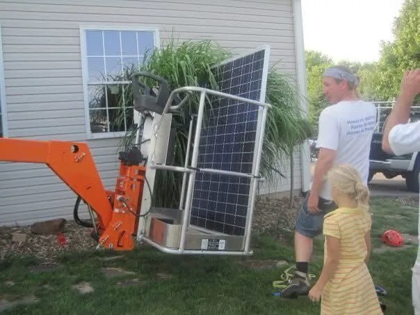 The first solar panel the company installed: a blue polycrystalline module in an orange lift cradle beside a house, one of the founders steadying it while a child watches