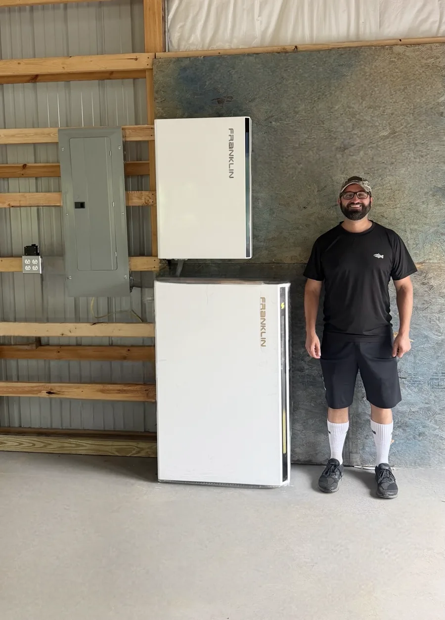A FranklinWH whole-house battery system mounted on a pole-barn wall beside a breaker panel, with a man standing next to it