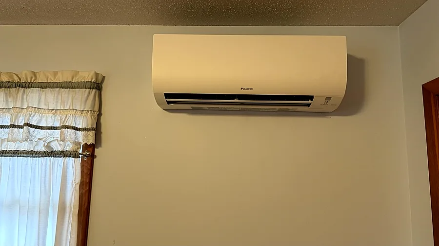 Wall-mounted ductless indoor head on a cream wall beside a curtained window