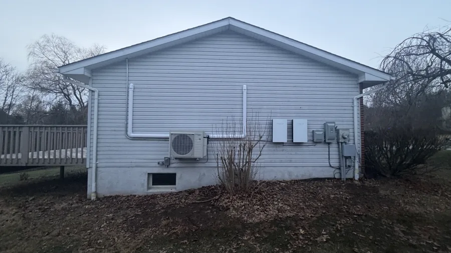 Outdoor unit on the side of a pale grey house with the line-set cover running up to the roofline